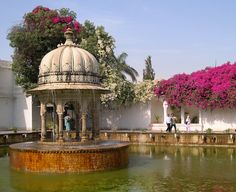 people are walking around in front of a fountain with pink flowers on the walls and trees