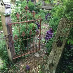 an iron gate in the middle of a garden with purple flowers and trees around it