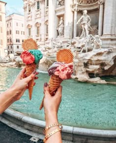 two people holding up ice cream cones in front of a fountain with statues on it