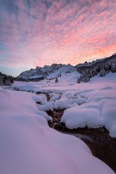 a stream running through a snow covered mountain valley under a pink sky with clouds in the distance