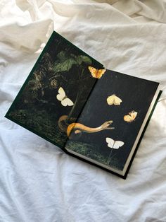 an open book with butterflies on it sitting on a white bed sheet in front of a window