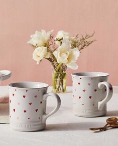 two coffee mugs with hearts on them sitting next to a vase filled with flowers