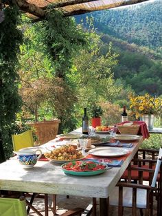 an outdoor dining table with plates and bowls on it, in front of a mountain view