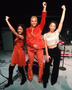 three women standing in front of a stage with their arms up and one woman wearing an orange suit