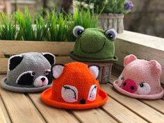 three crocheted hats with different animals on them sitting on a wooden table next to plants