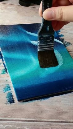 a person holding a paint brush over a piece of art on a wooden table with blue and green colors