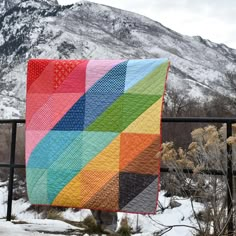 a multicolored quilt hanging on a fence in front of a snow covered mountain