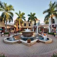 an outdoor fountain surrounded by palm trees in a shopping center with people sitting around it