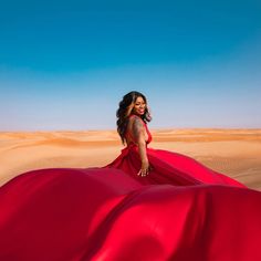 a woman in a long red dress standing on top of a sand dune with blue sky behind her