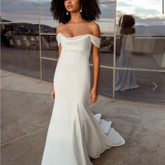 a woman standing in front of a glass wall wearing a white wedding dress with an off the shoulder neckline