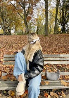 a woman sitting on top of a wooden bench in a park filled with leaves and trees