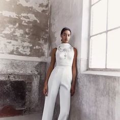 a woman standing in front of a window wearing a white jumpsuit and high heels