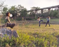 three young women playing frisbee in an open field with a bridge in the background