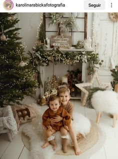 two young children sitting on a rug in front of a christmas tree with lights and garlands