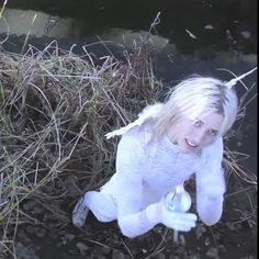 a woman with blonde hair sitting on the ground next to a body of water and grass