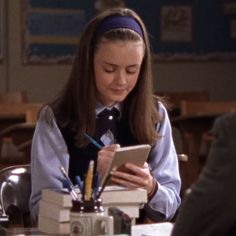 a woman sitting at a table in front of a stack of books and writing on a clipboard