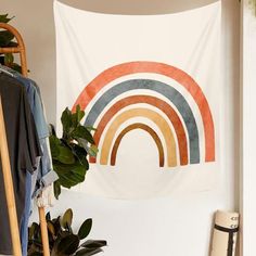 a wall hanging with a rainbow print on it next to a potted plant and clothes rack