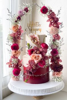 a three tiered cake decorated with flowers and greenery on a table in front of a window