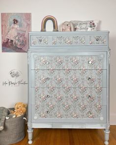 an old dresser painted in blue and white with flowers on the front, sitting next to a teddy bear