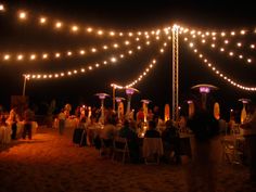 people sitting at tables under string lights on the beach