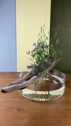 a glass bowl filled with water and flowers on top of a wooden table next to a piece of driftwood