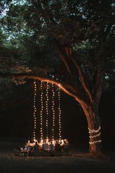 a group of people sitting around a table under a tree with lights hanging from it