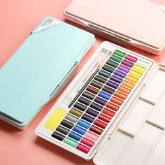 an assortment of watercolors in a case on a pink surface with one open and the other closed