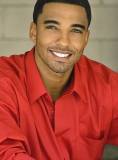 a man in a red shirt sitting on the ground smiling at the camera with his arms crossed