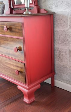 a red dresser with two drawers and a mirror on it's top, in front of a brick wall