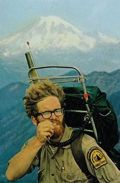 a man sitting on top of a mountain next to a backpack and looking at the camera