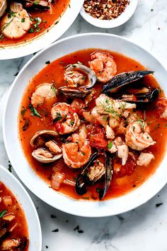 three bowls filled with seafood and clams in tomato sauce on top of a marble table