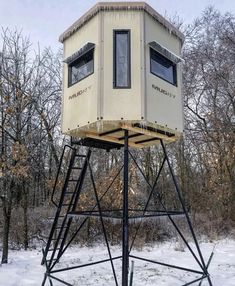 a tall tower with two windows in the middle of a snow covered field next to trees