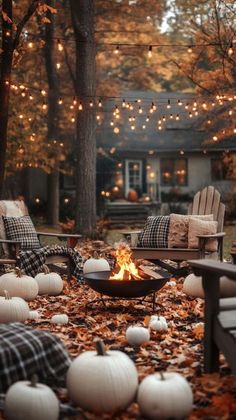 a fire pit surrounded by fall leaves and pumpkins in front of a house with string lights