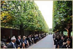 many people are sitting on benches in front of trees and bushes, while one person is walking down the sidewalk