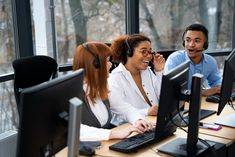 three people sitting in front of computer monitors talking on headsets and smiling at each other