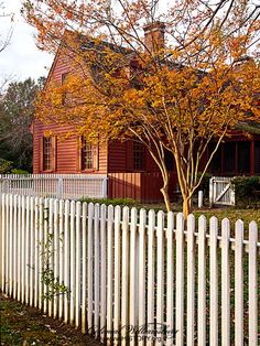 a white picket fence in front of a red house and tree with orange leaves on it