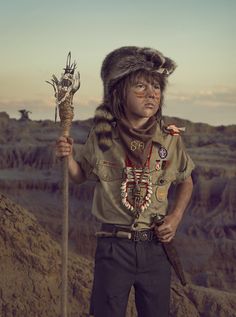 a man wearing a fur hat and holding a stick in his hand while standing on top of a mountain