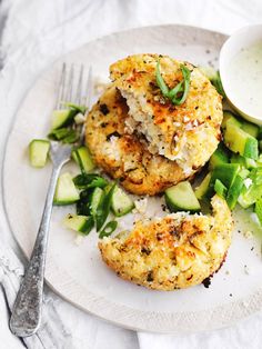 a white plate topped with crab cakes next to cucumbers and a bowl of sauce