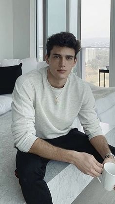 a man sitting on top of a bed next to a window holding a coffee cup