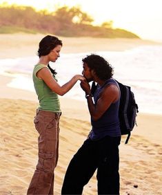 two people standing on the beach with one touching the other's ear and looking at each other