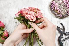 someone is arranging flowers on a table with scissors and wire work around them, including pink roses