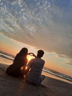 a man and woman sitting on the beach at sunset making a heart shape with their hands