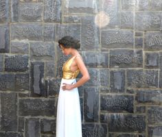 a woman standing in front of a stone wall wearing a white and gold colored dress