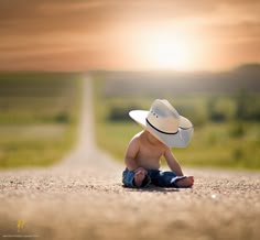 a baby sitting on the ground wearing a cowboy hat and looking at the camera with an empty road in the background