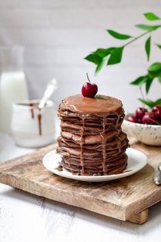 a stack of chocolate pancakes sitting on top of a wooden cutting board next to a bowl of cherries