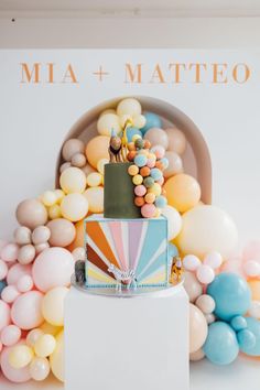 a colorful cake sitting on top of a white table next to balloons and a sign