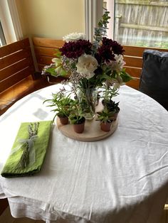 a white table topped with flowers and potted plants next to a green napkin on top of a wooden chair