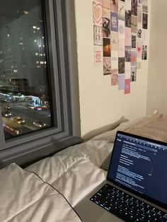 an open laptop computer sitting on top of a bed next to a window with city lights in the background