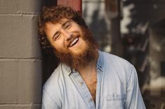 a bearded man leaning against a brick wall and smiling at the camera with his eyes closed