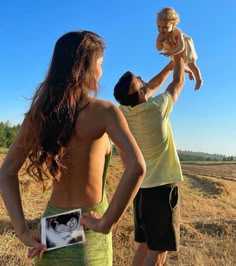 a man holding a baby up to his face while standing next to a woman in a field